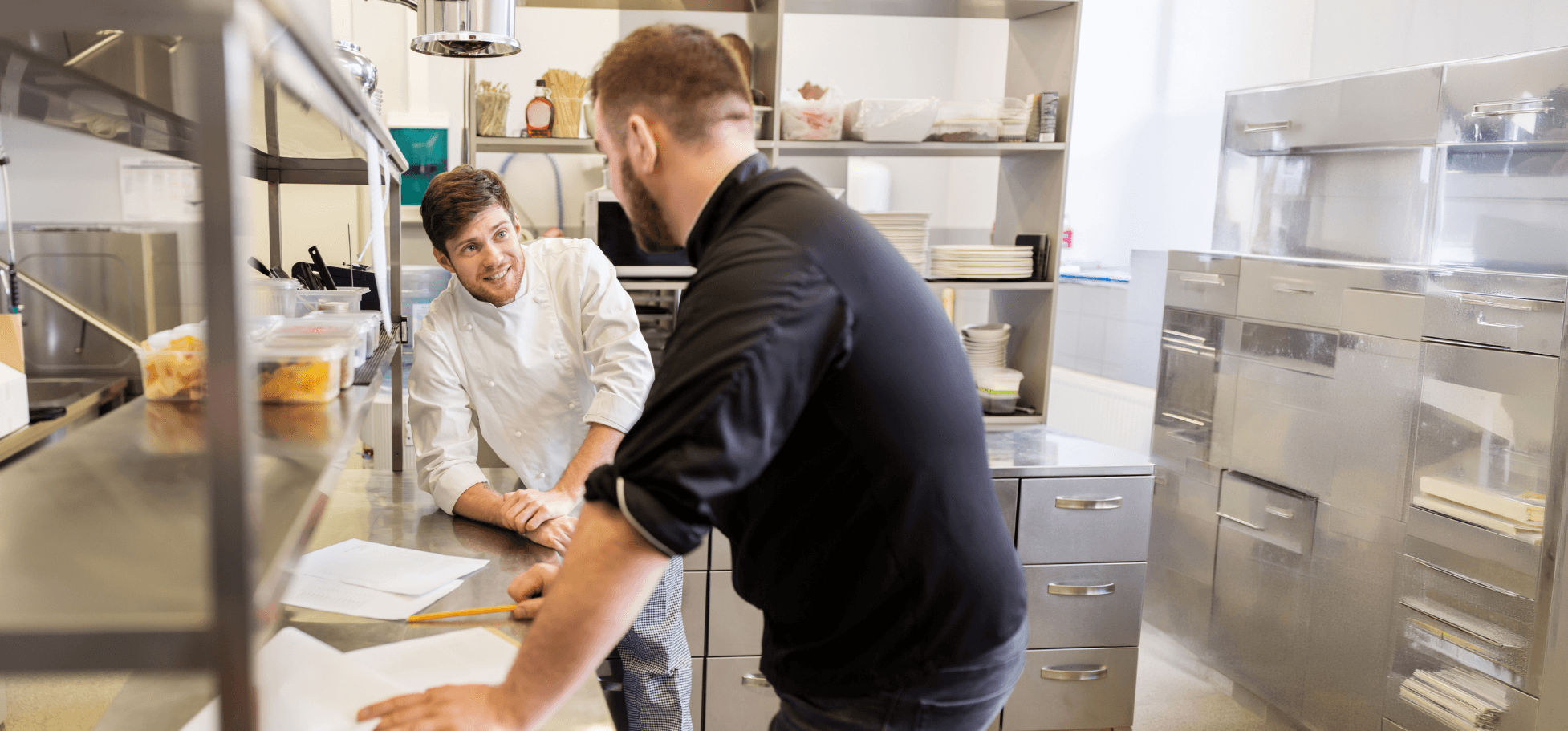 Chefs discussing paperwork inside a professional kitchen.